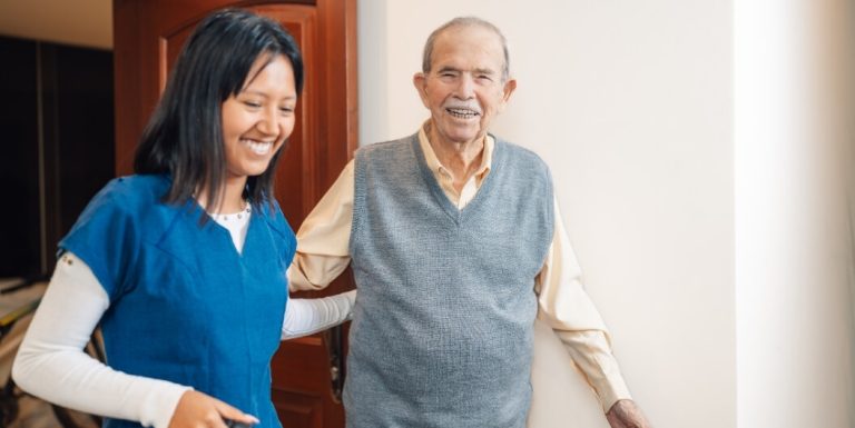 Smiling Elderly Man Standing Beside a Female Nurse in a Bright Hallway at an Assisted Living Facility
