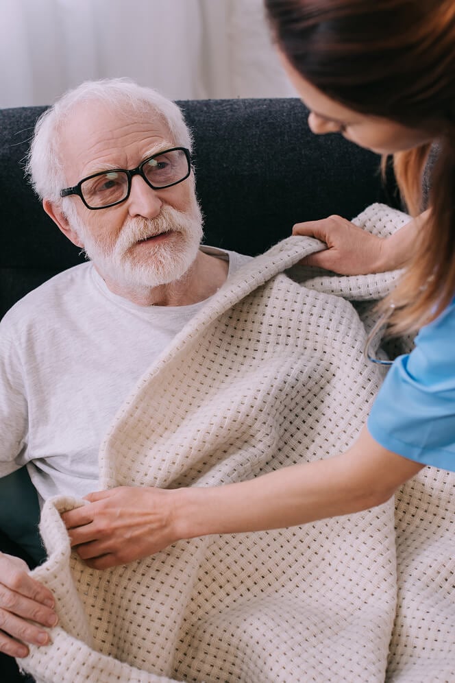 Female Nurse Tucks Warm Blanket Around Elderly Man on Sofa Female Nurse Tucks Warm Blanket Around Elderly Man on Sofa